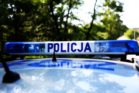 Sygnały świetlne i dźwiękowe na dachu pojazdu. Na białym tle niebieski napis Policja, po lewej i po prawej stronie światła koloru niebieskiego. W tle drzewa.
