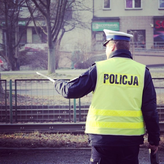 Umundurowany policjant ruchu drogowego, w kamizelce odblaskowej z napisem Policja, stojący tyłem. W lewej ręce trzyma tarcze sygnalizacyjną. W tle zabudowania.
