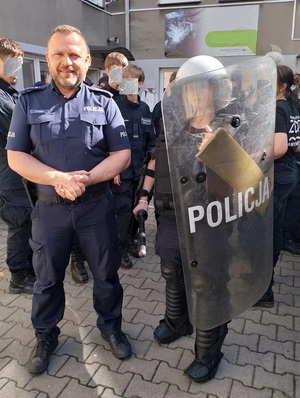 Po lewej stronie umundurowany policjant, po prawej osoba ubrana w elementy policyjnego stroju, trzymająca w ręku tarcze z napisem Policja. W tle uczniowie.