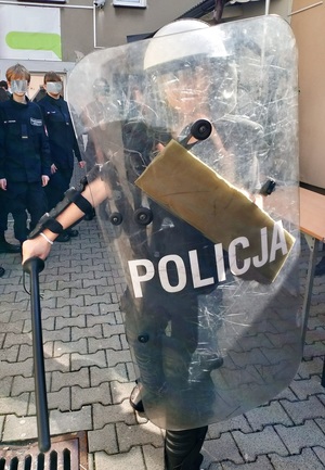 Na zdjęciu osoba ubrana w elementy stroju policyjnego, trzymająca w ręku tarcze z napisem Policja.