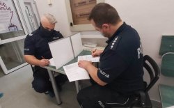 Dwóch umundurowanych policjantów z maseczkami na twarzy zasłaniającymi usta i nos, którzy siedzą przy stoliku na którym znajduje się przegroda i wypełniają dokumenjty.