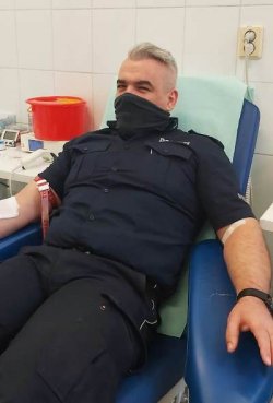 Umundurowany funkcjonariusz Policji, z maseczką na twarzy zasłaniającą usta i nos, siedzący na łóżku szpitalnym, oddający krew.