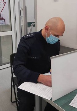 Umundurowany policjant z maseczką założoną na usta i nos, siedzący przy stoliku, wypełniający dokumenty.