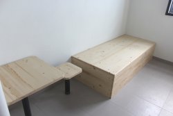 Pomieszczenie dla osób zatrzymanych. Po prawej stronie drewniane łóżko, obok drewniany solik i krzesło.
