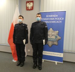 Dwóch nowo przyjętych do służby umundurowanych policjantów, stojących przodem, trzymających w ręku akty ślubowania. Po lewej stronie flaga Polski, po prawej stroni policyjny baner, a w tle Godło Polski.