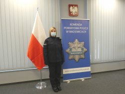 Nowo przyjęta policjantka, umundurowana. Po lewej stronie znajduje się Flaga Polski, po prawej baner z napisem Komenda Powiatowa Policji w Wadowicach z odznaką policyjną, a także Godło Polski.
