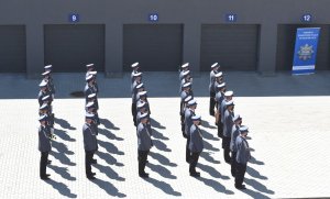 Zdjęcie z lotu ptaka. Policjantki i policjanci w umundurowaniu galowym, stojący w pięciu szeregach na placu komendy.