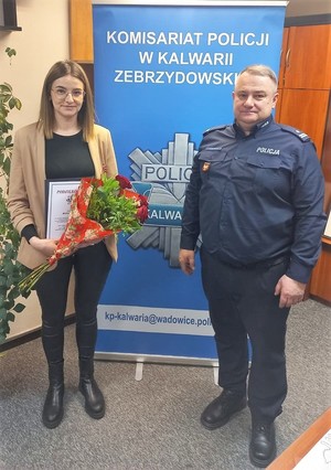 Na zdjęciu po lewej stronie kobieta w stroju cywilnym trzymająca w ręku kwiaty i list gratulacyjny, po prawej umundurowany policjant. W tle policyjny baner z napisem Komisariat Policji w kalwarii Zebrzydowskiej.