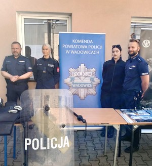 Pośrodku policyjny baner n napisem Komenda powiatowa Policji w Wadowicach. Po prawej i po lewej stronie umundurowany policjant i uczennice klas mundurowych.