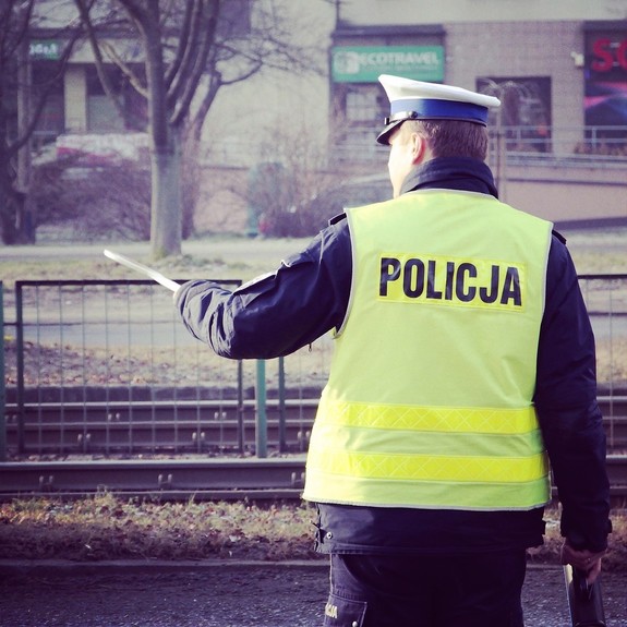 Umundurowany policjant ruchu drogowego w kamizelce odblaskowej z napisem Policja, stojący tyłem, w lewej ręce trzyma tarcze sygnalizacyjną. W tle zabudowania.