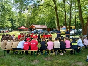 Grupa osób siedzących tyłem na ławkach, przed nimi umundurowana policjantka i policjant. W tle namiot, drewniany budynek, grupa osób siedzących na ławkach, samochody i drzewa.