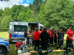 Grupa osób siedzących tyłem, przed nimi umundurowany policjant ruchu drogowego stojący w oznakowanym radiowozie. W tle drzewa.