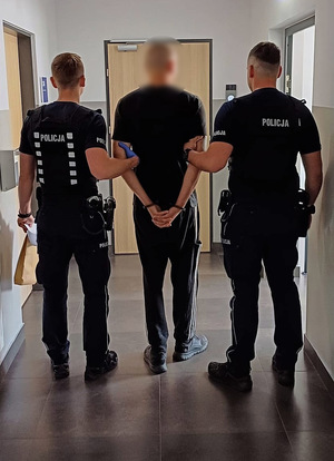 Zatrzymany mężczyzna ubrany na ciemno, na rękach z tyłu ma założone kajdanki. Po prawej i po lewej stronie stoją umundurowani policjanci.