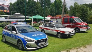 Na zdjęciu po lewej stronie oznakowany radiowóz, dalej dwa pojazdy straży pożarnej. W tle rozstawione parasole i ławki.