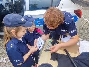 Na zdjęciu umundurowana policjantka, obok której stoją dwie małe dziewczynki ubrane w strój policjanta.  Policjantka przekazuje jednej z ich odbite linie papilarne.