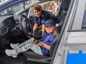 Umundurowana policjantka na miejscu pasażera w oznakowanym radiowozie, na miejscu kierowcy siedzi mała dziewczynka ubrana w koszulę i czapkę policyjną, w rękach trzyma kajdanki i zabawkowy pistolet.