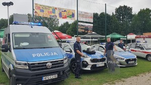 Na zdjęciu trzy oznakowane radiowozy oraz trzech umundurowanych policjantów, Jeden z nich trzyma tarcze policyjną.