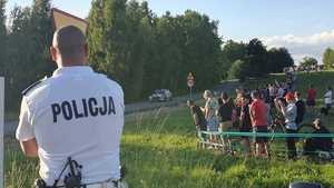 Na zdjęciu policjant w białej koszulce z napisem Policja stojący tyłem, po prawej stronie grupa kibiców stojących na trawie. W tle jadący po drodz\e samochód wyścigowy.