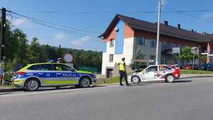 Po lewej oznakowany radiowóz, ma skrzyżowaniu policjant, który kieruje ruchem. Po prawej samochód wyścigowy. W tle budynek.