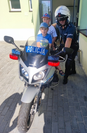 Na policyjnym motocyklu siedzą dwaj chłopcy. Obok stoi umundurowany policjant.