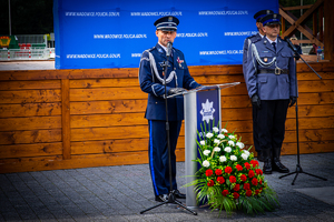 Komendanta Powiatowy Policji w Wadowicach stojący i przemawiający przy mównicy, przed którą  jest kosz z biało czerwonymi kwiatami. W tle dwóch policjantów.