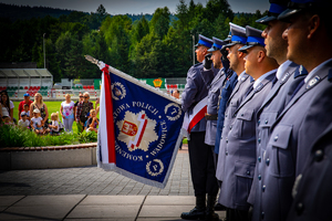 Stojący w szeregu policjanci w strojach galowych. Pierwszy od lewej strony trzyma sztandar.