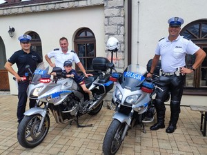 Na zdjęciu trzech policjantów ruchu drogowego oraz dwa policyjne motocykle, na jednym siedzi mały chłopiec ubrany w capkę i koszulę policyjna.