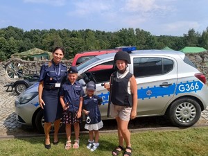 Po lewej stronie umundurowana policjantka i trzy dziewczynki ubrane w elementy policyjnego munduru. Stoją na tle radiowozu.