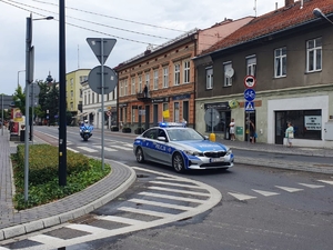 Jadący oznakowany radiowóz, a za  nim policyjny motocykl. W tle budynki.