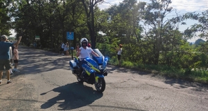 Policjant jadący na motocyklu. W tle drzewa i kibice.