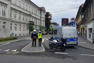 W rejonie skrzyżowania samochód policyjny i motocykl oraz stojący tyłem policjant i policjantka. W tle zabudowania.