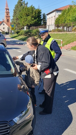 Policjant WRD stoi przy zatrzymanym samochodzie, uczeń szkoły wraz z wychowawcą klasy wręczają kierowcy samochodu pojemnik z cytrynami.