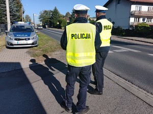 Dwóch policjantów ruchu drogowego w kamizelkach odblaskowych stojących tyłem przy drodze. W tle radiowóz.