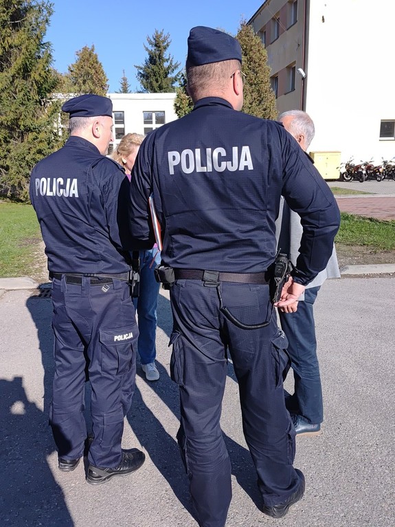 Na tle budynku szkoły dwóch policjantów stojących tyłem, Przed nimi kobieta i mężczyzna.