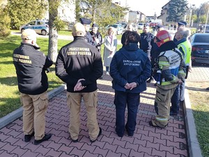 Na zdjęciu umundurowani policjanci i strażacy oraz przedstawiciele zarządzenia kryzysowego. W tle  budynki, ludzie i samochody.