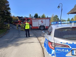 Po prawej stronie radiowóz  i stojące powyżej wozy strażackie. Na drodze stojący tyłem umundurowany policjant i grupa strażaków.