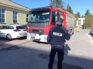 Policjant stojący tyłem na tle wozu strażackiego. W tle strażak i przewodnik z psem tropiącym.