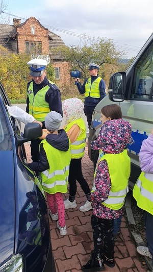 Na zdjęciu widać dwóch umundurowanych policjantów ruchu drogowego oraz grupę dzieci w kamizelkach odblaskowych.