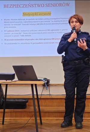 Umundurowana policjantka trzymająca w ręku mikrofon. Obok niej stolik na którym jest komputer, a za nią ekran, na którym jest wyświetlana prezentacja odnośnie niebezpieczeństwa seniorów.