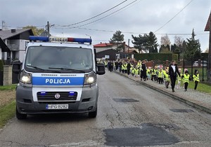 Po lewej stronie radiowóz, po prawej grupa dzieci w kamizelkach odblaskowych idących chodnikiem.