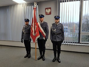 Trzech umundurowanych policjantów, z których jeden trzyma sztandar. W tle Godło Polski.