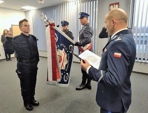 Nowo przyjęty policjant ślubujący na sztandar. Po prawej stronie Komendant Powiatowy Policji w Wadowicach, pośrodku trzech umundurowanych policjantów, z których jeden trzyma sztandar.