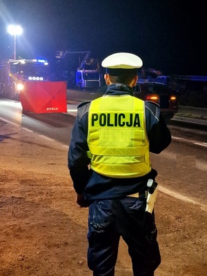 Miejsce śmiertelnego wypadku drogowego. Na zdjęciu widać stojącego tyłem umundurowanego policjanta. W tle rozłożony parawan, wóz strażacki oraz samochód cywilny.