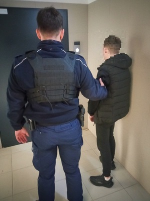 Umundurowany policjant trzymający za rękę zatrzymanego mężczyznę, który jest ubrany na czarno.