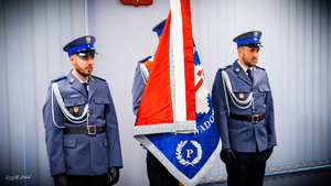 Poczet sztandarowy. Trzech umundurowanych policjantów, z których funkcjonariusz stojący w środku trzyma sztandar wadowickiej komendy.