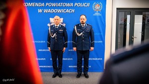 Na tle policyjnego baneru stoi dwóch funkcjonariuszy Policji w mundurach galowych.