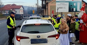 Na zdjęciu po lewej stronie stoi umundurowany policjant obok samochodu osobowego koloru białego, a po prawej stronie grupa dzieci i Mikołaj.