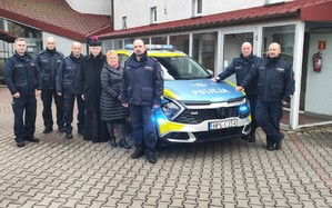 Na tle oznakowanego radiowozu stoi sześciu umundurowanych policjantów, kobieta w stroju cywilnym oraz ksiądz.