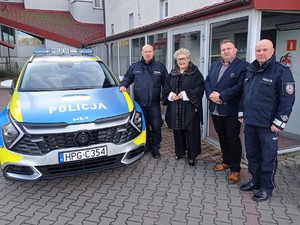 Na zdjęciu dwóch umundurowanych policjantów oraz kobieta i mężczyzna  stroju cywilnym, a także oznakowany radiowóz.