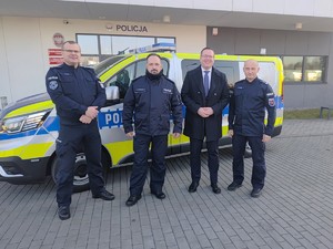 Na tle oznakowanego radiowozu stoi trzech umundurowanych policjantów oraz  mężczyzna w stroju cywilnym. W tle budynek komendy.